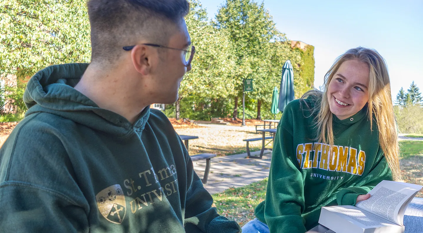 A female student wearing a St. Thomas hoodie holds open a book while looking at a male classmate outside on campus