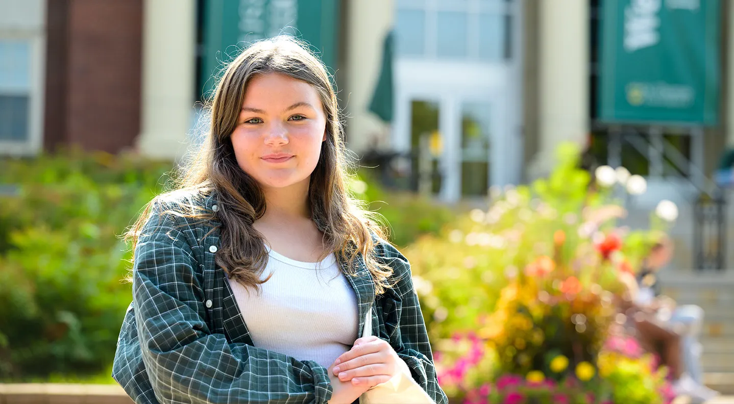 A female student clutches a tote bag and smiles at the camera in front of George Martin Hall