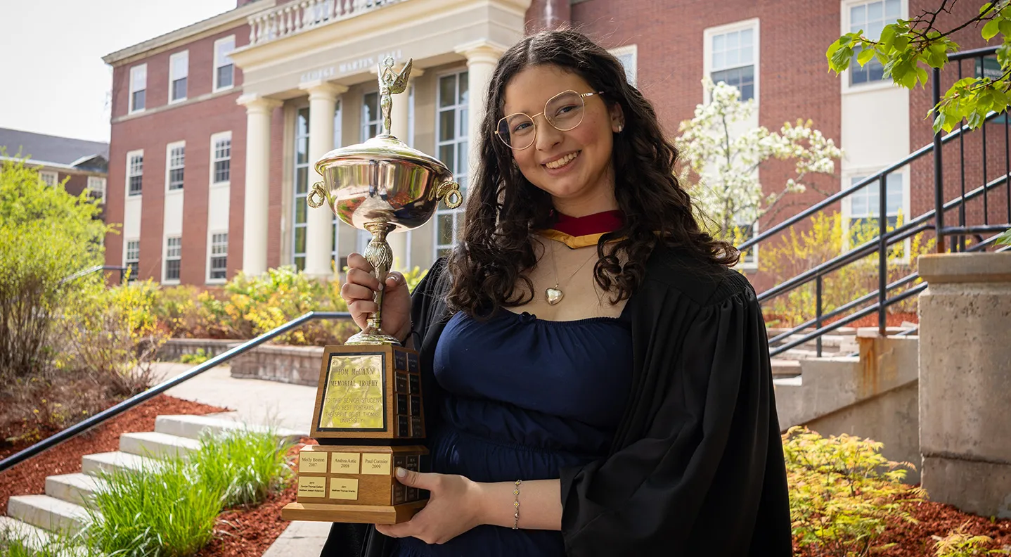 Ana Lucia Pavon stands beneath a tree on campus, wearing her grad gown and holding the Tom McCann Memorial Trophy