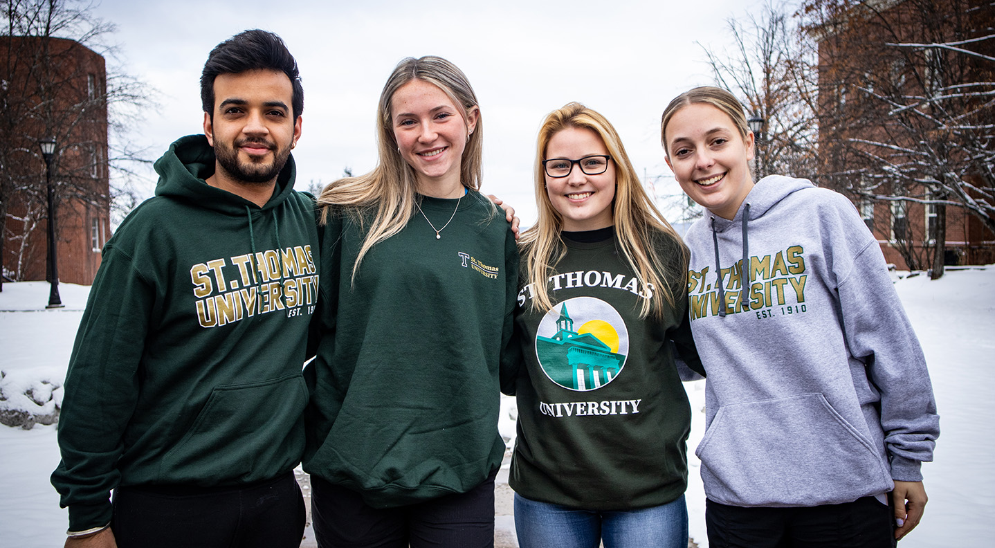 A group of students—one male, three female—stand arm in arm on a snowy day on campus