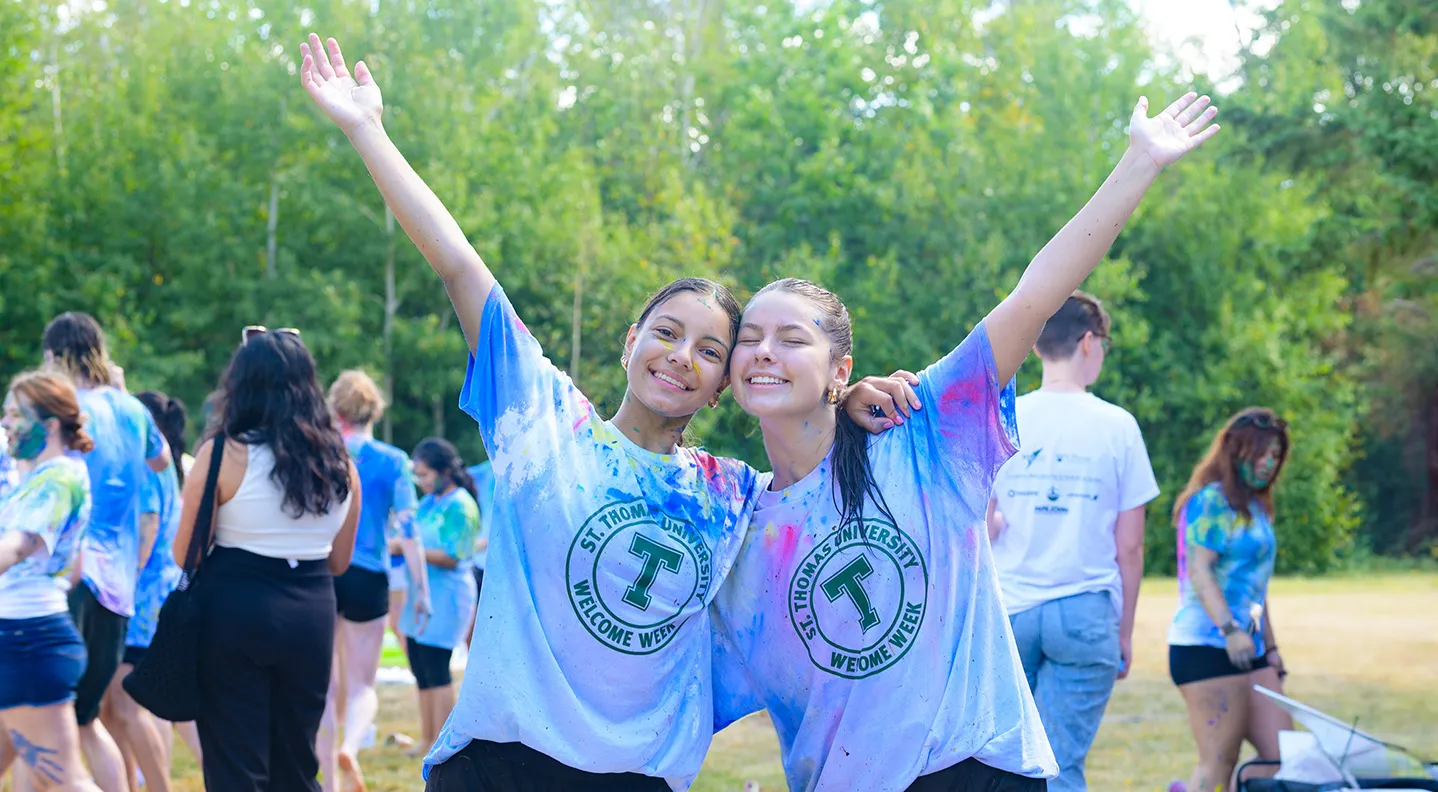Two female student stand smiling with their hands in the air, covered in paint following the annual Welcome Week Paint Fight