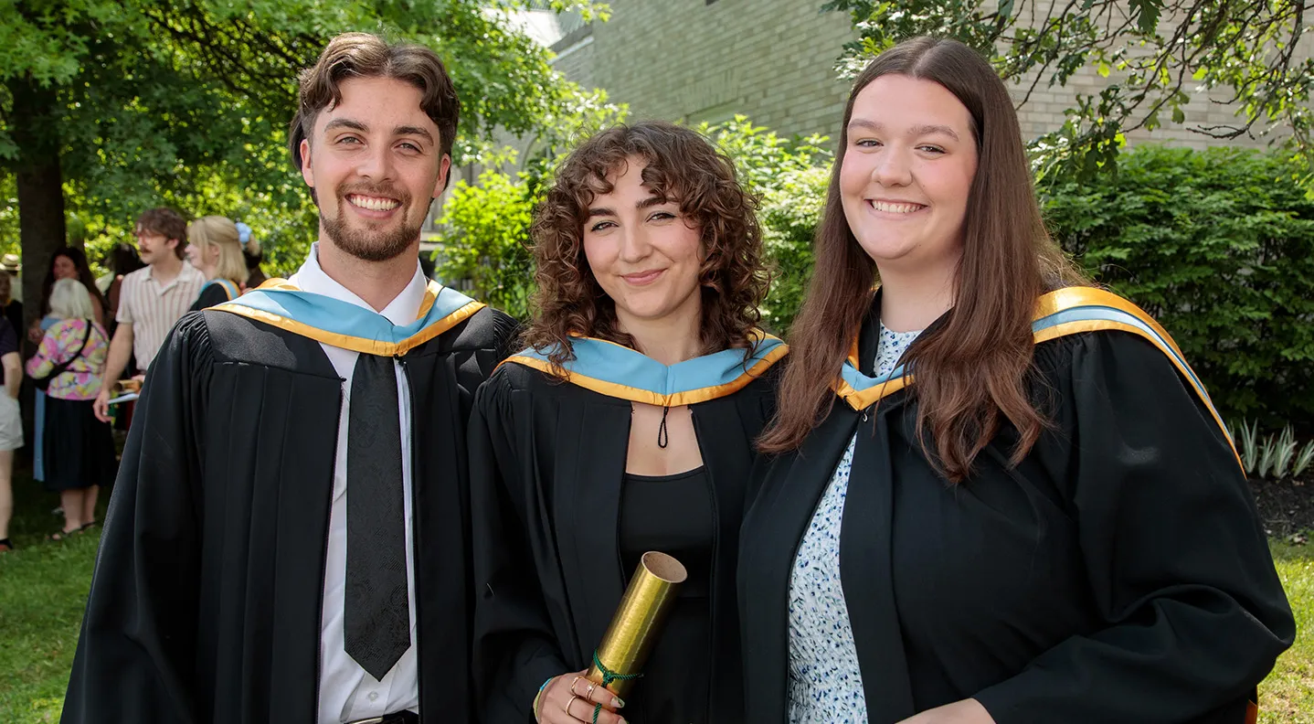 Three graduates stand together smiling outside the Fredericton Playhouse
