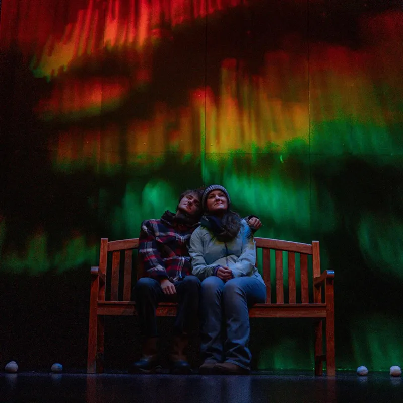 Two individuals sitting on a bench with the reflection of the Northern Lights behind them during a production of Almost, Maine