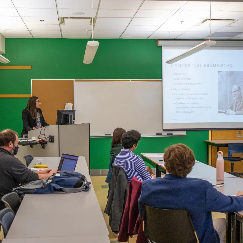 A student presenting their research in class with a professor and a group of students in attendance