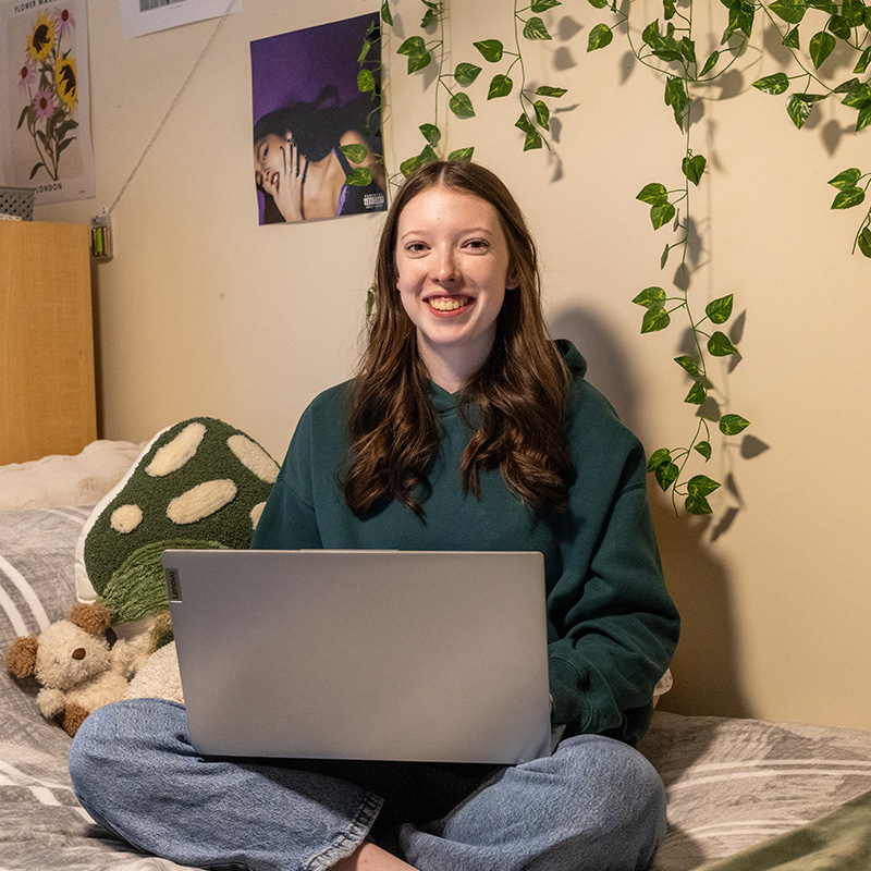 A female student sits cross-legged on her bed with her laptop in her lap