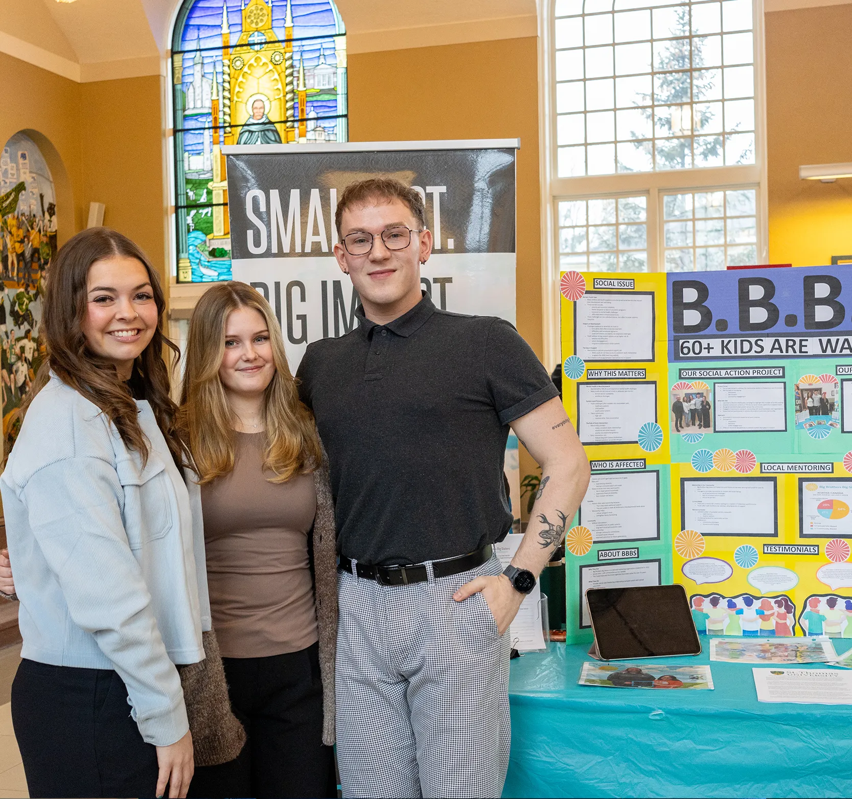 Two female students and one male student standing similing in front of a banner for the Big Brothers Big Sisters of Fredericton and Oromocto