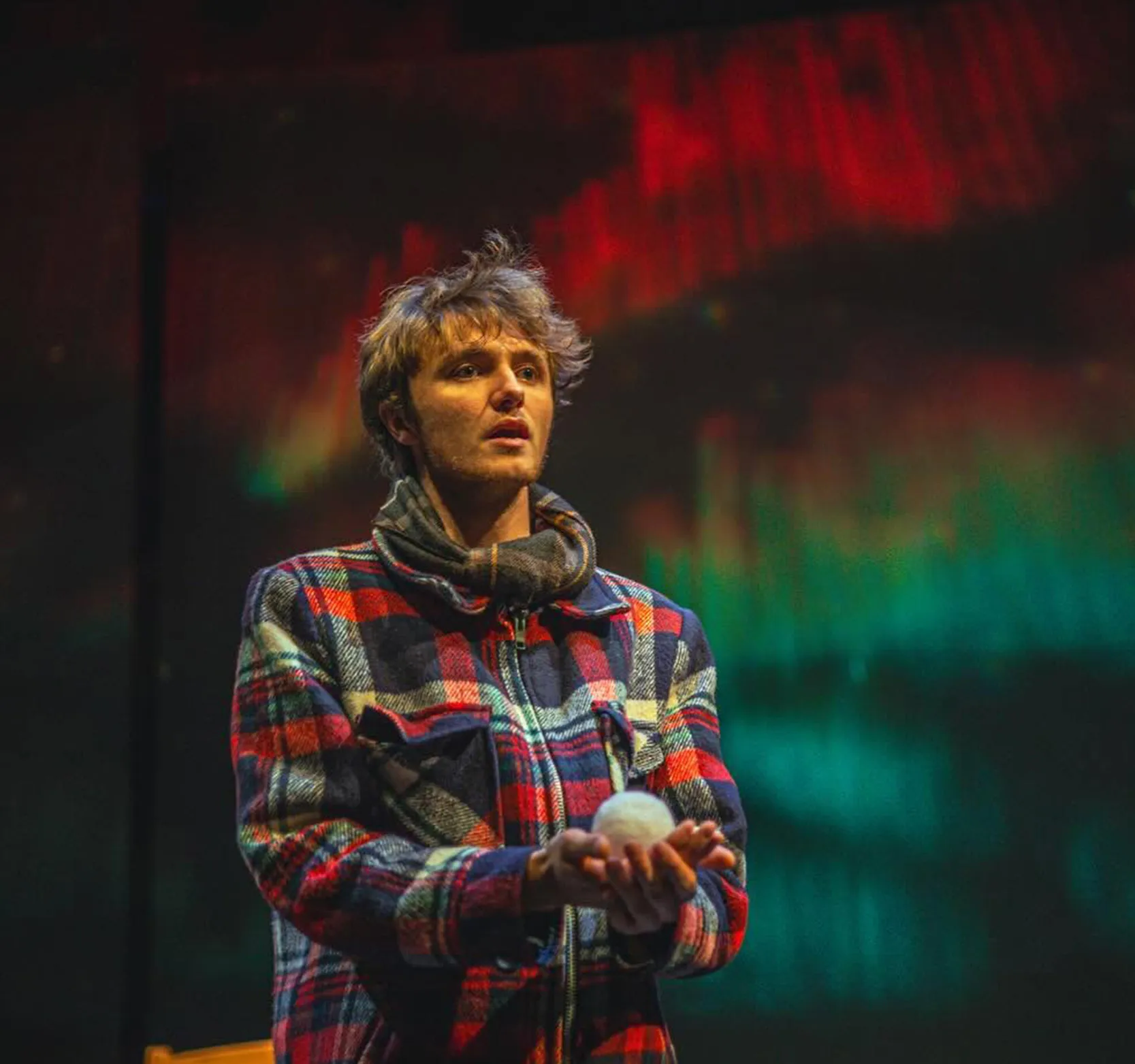 A male actor in a plaid coat holds a fake snowball in front of the northern lights in the Black Box Theatre