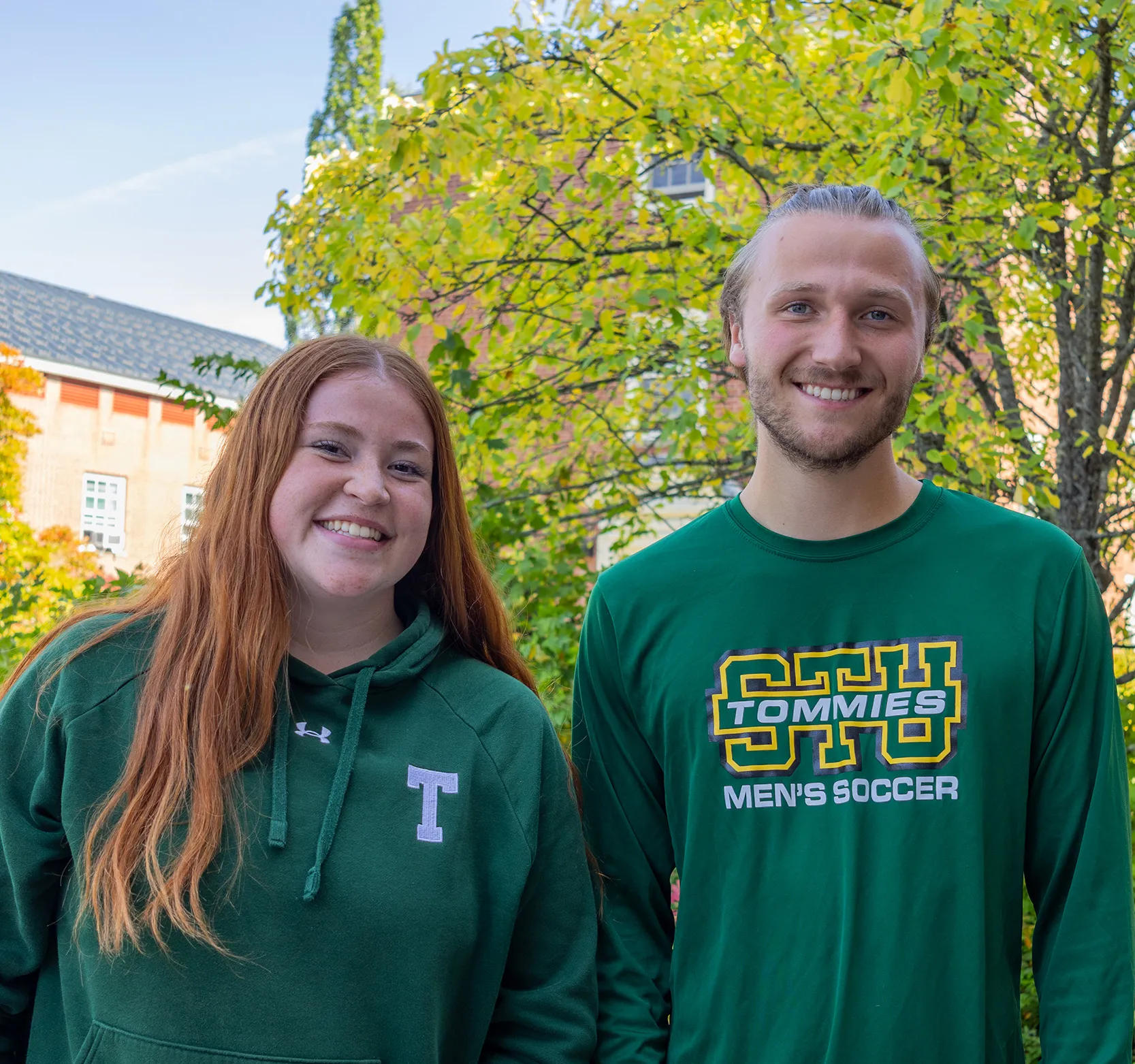 A female student with red hair and a green hoodie stands beside a male student with blonde hair wearing a STU Tommies Soccer shirt