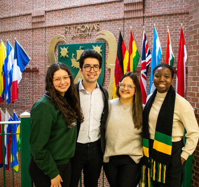 Four international students smiling next to wall of flags in James Dunn Hall at 鶹