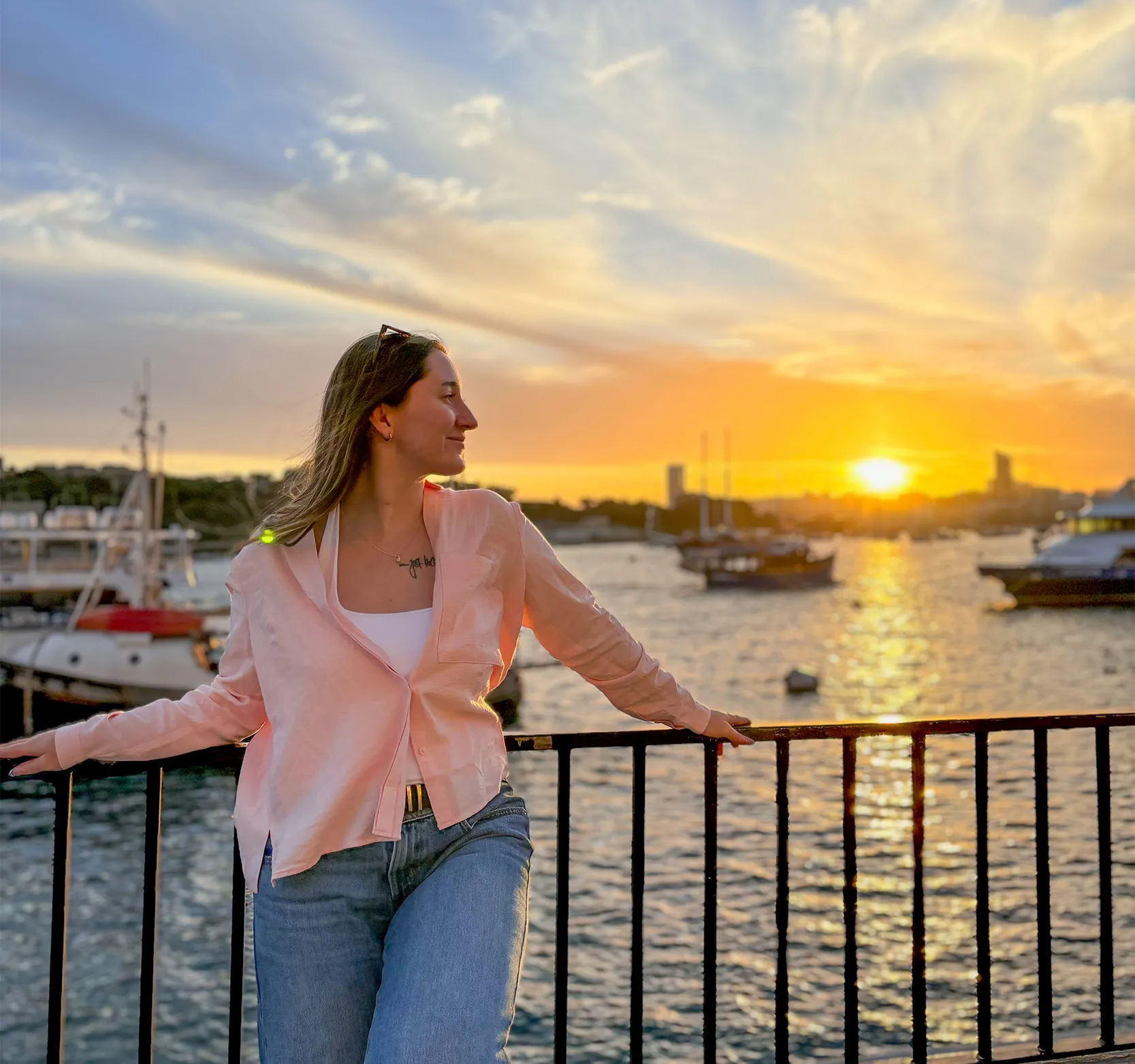 Kamylle leaning against a waterfront railing in Malta, looking toward a golden sunset over the harbour, with boats and buildings in the background.