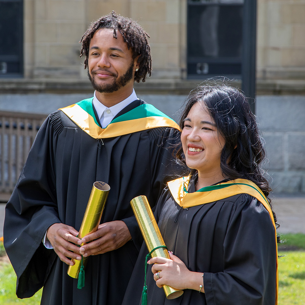 Two Master of Social Work grads stand together in their gowns holding their degrees