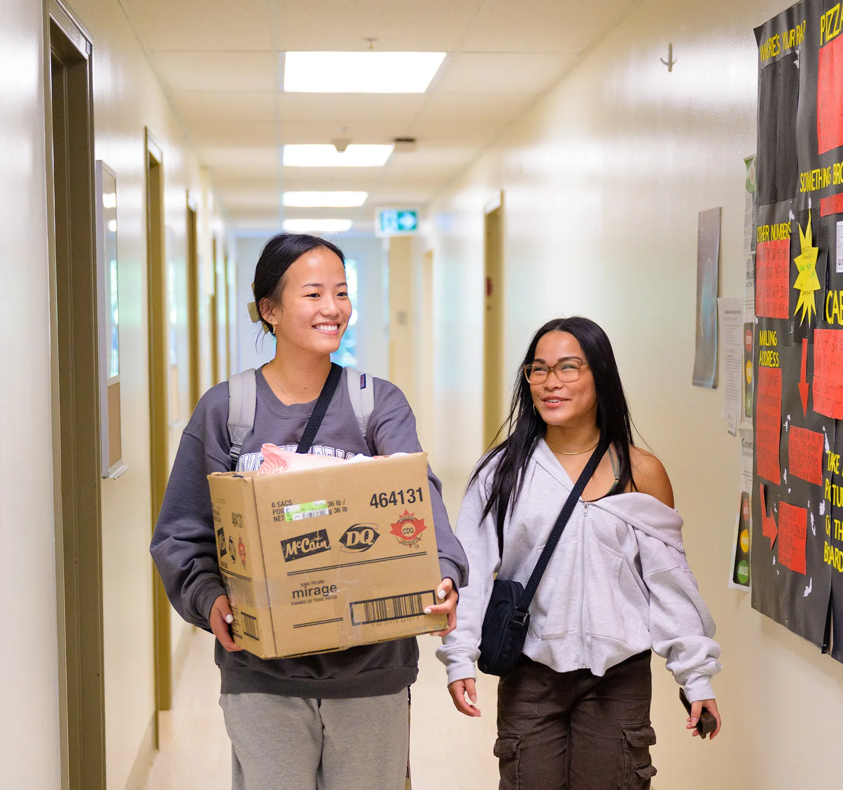 A female student walks down the hall holding a cardboard box alongside her mom, who is smiling at her.