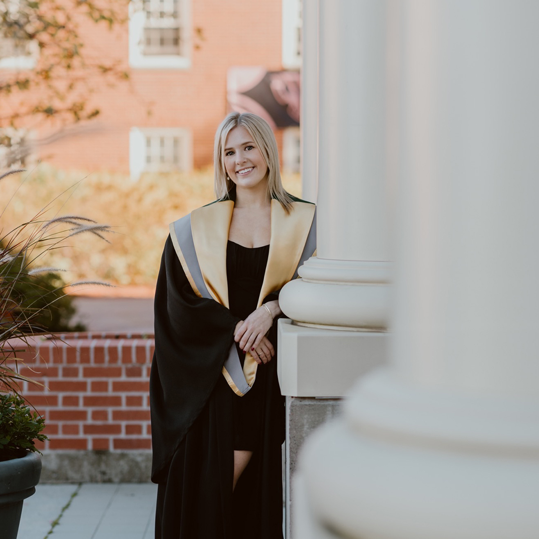 Avery White in grad gown and BSW hood leaning against George Martin Hall